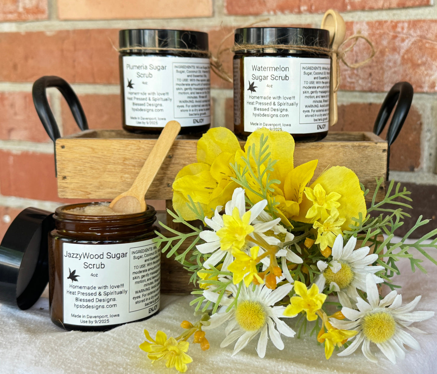 Various jars of Sugar Scrub with a wooden box and flowers on a brick background