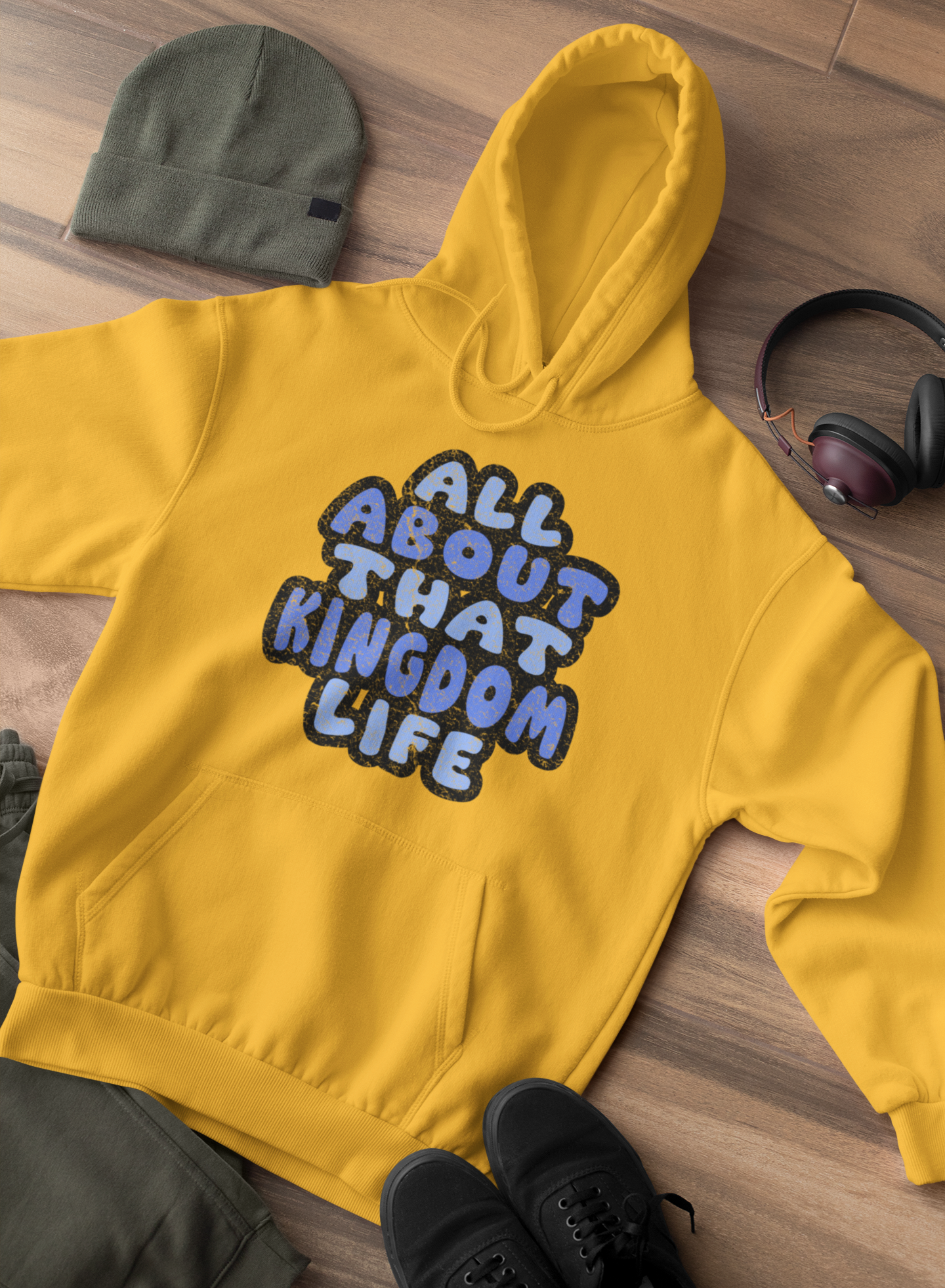 Gold hoodie with distressed text on a wooden floor, surrounded by a green beanie, black shoes, and headphones.