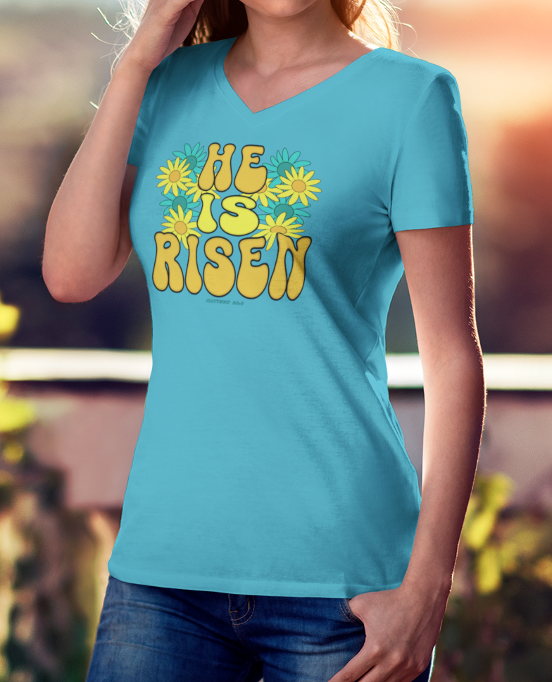 Woman wearing a turquoise blue v-neck t-shirt with 'He is Risen' text, standing outdoors.
