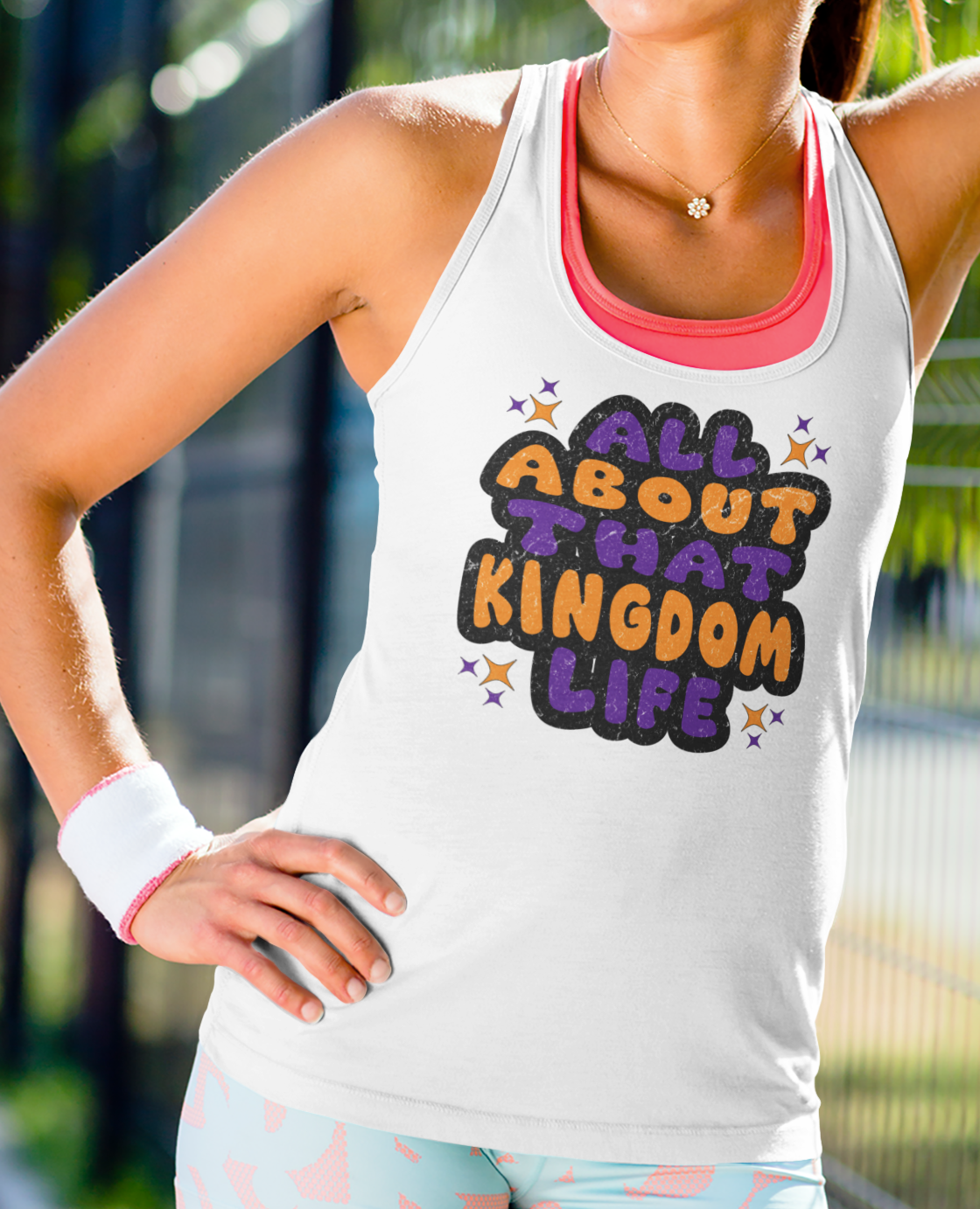 Woman wearing a white racerback tank top with 'All About That Kingdom Life' distressed text outdoors.